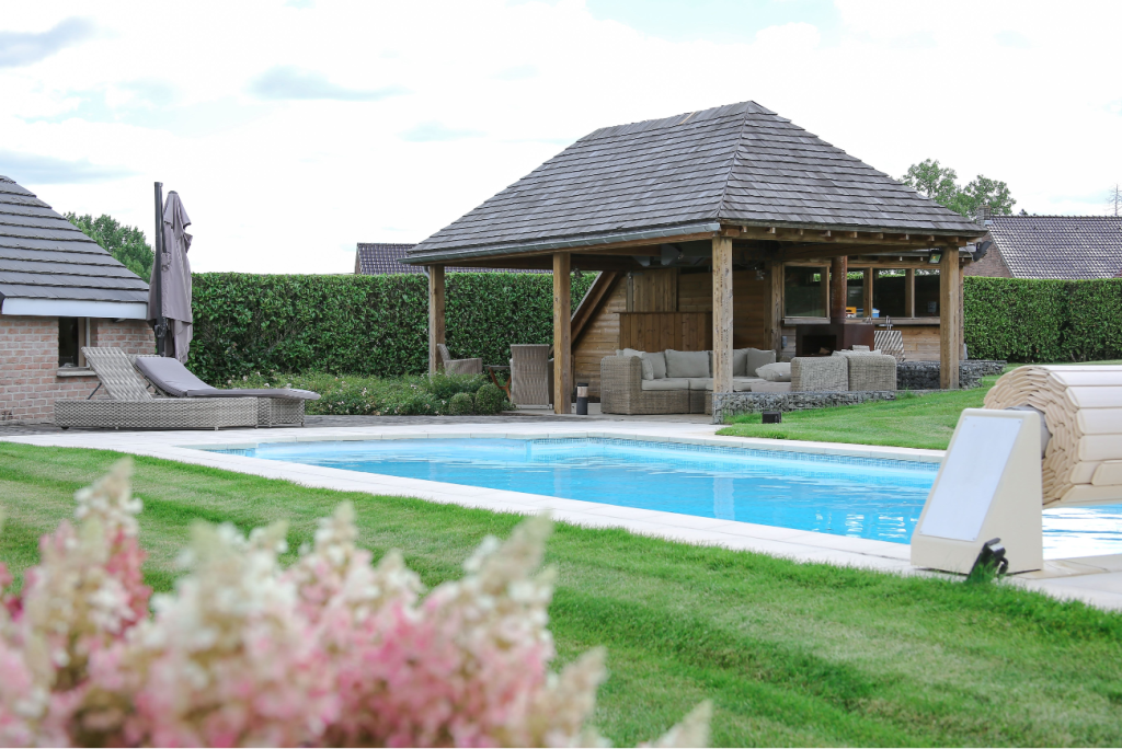 Pavillon en bois à toiture d’ardoise au bord d’une piscine, entouré d’une pelouse et d’une haie