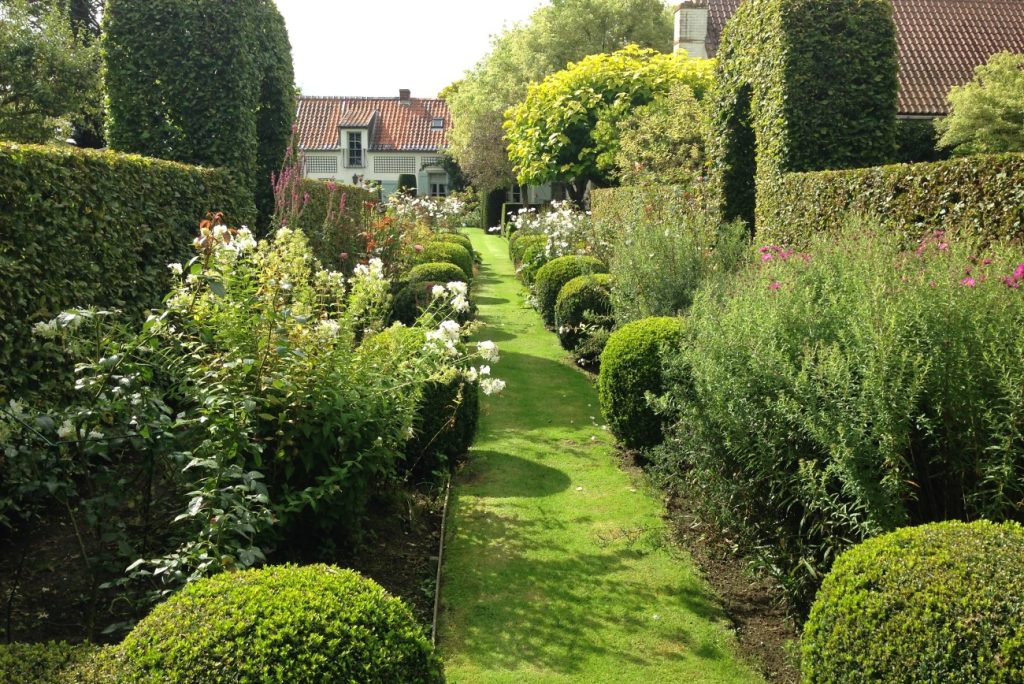 Allée engazonnée bordée de massifs fleuris et de boules de buis, menant à une maison