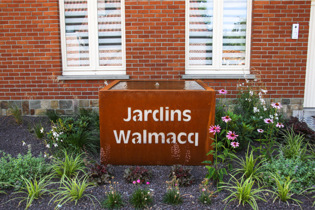 Point d’eau en acier corten gravé « Jardins Walmacq » au milieu de massifs fleuris