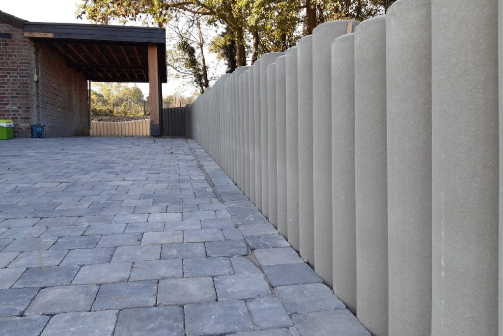 Terrasse en pavés gris bordée de palissades en béton, le long d’un abri en bois